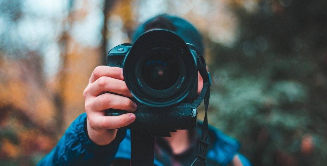 Photo of someone taking a photograph. Image looks into the lens of the camera, with autumnal trees behind.