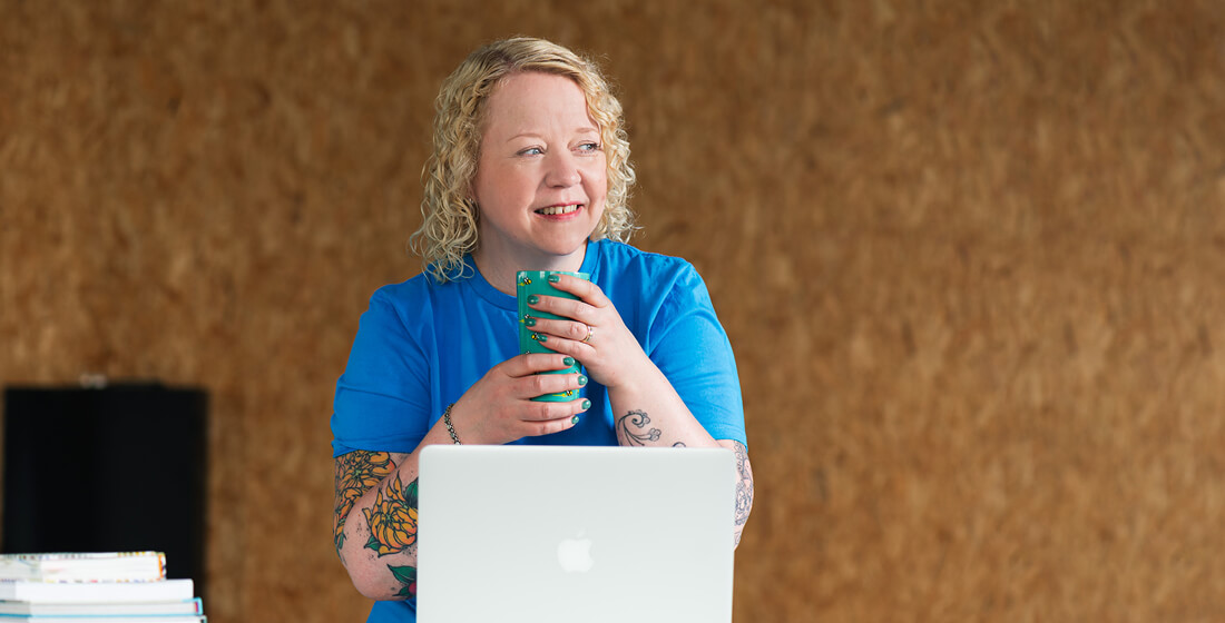 Helen Bee from B Double E working at a laptop, holding a mug and smiling.
