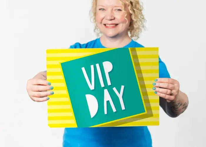 Helen Bee from B Double E holding a poster that says 'VIP Day' on a striped yellow background.