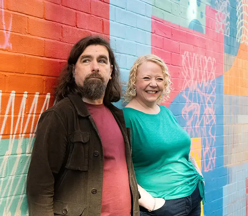 Rob Bee and Helen Bee from B Double E stood smiling, leaning against a brightly coloured graffiti wall.