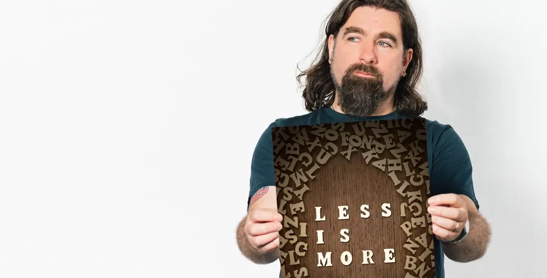 Rob Bee from B Double E holding a A3 card that says 'less is more'. He's stood against a plain pale background wearing a dark blue t-shirt.
