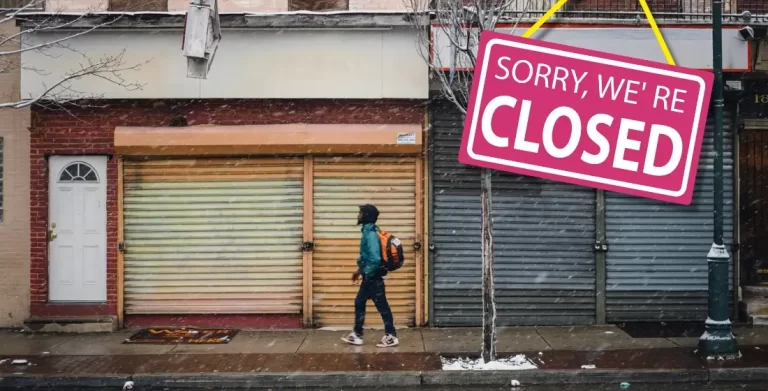 Metal shutters on shops with a 'sorry we are closed' graphic. There is a man in a hoody wearing a rucksack walking past the closed shops. Image from Unsplash by Dyana Wing So.